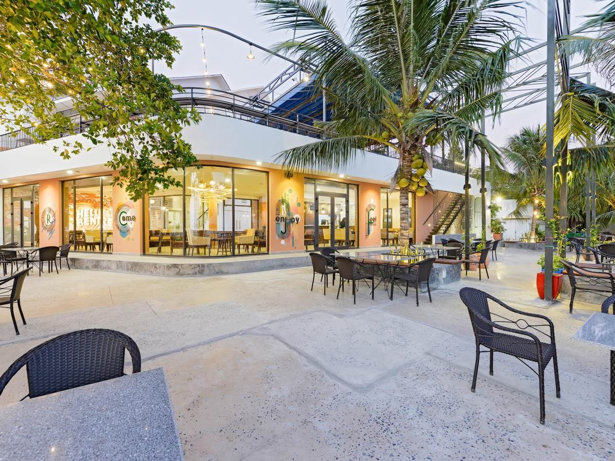 Open-air caf&eacute; terrace with dark rattan chairs under palms beside a glass-fronted building