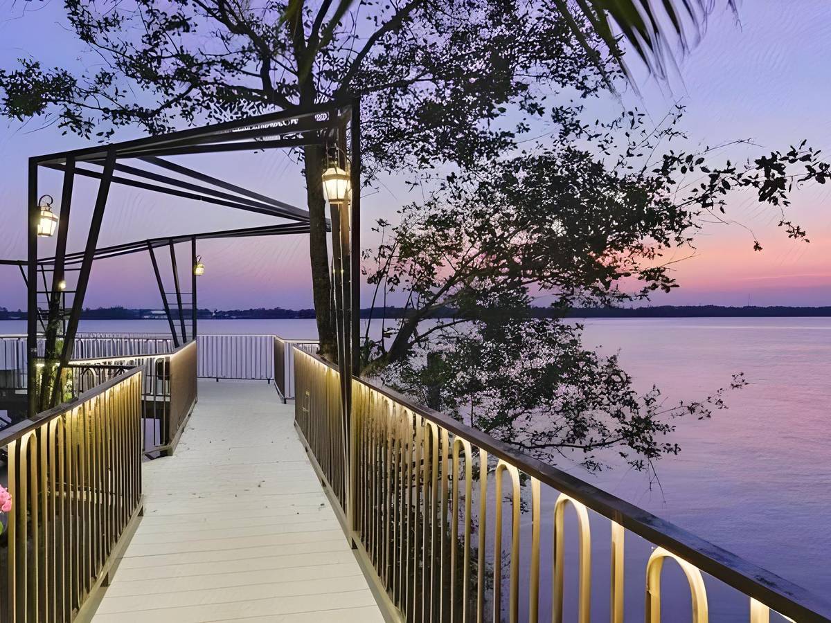 Long walkway with lit railings running out over a calm river at dusk under a steel frame