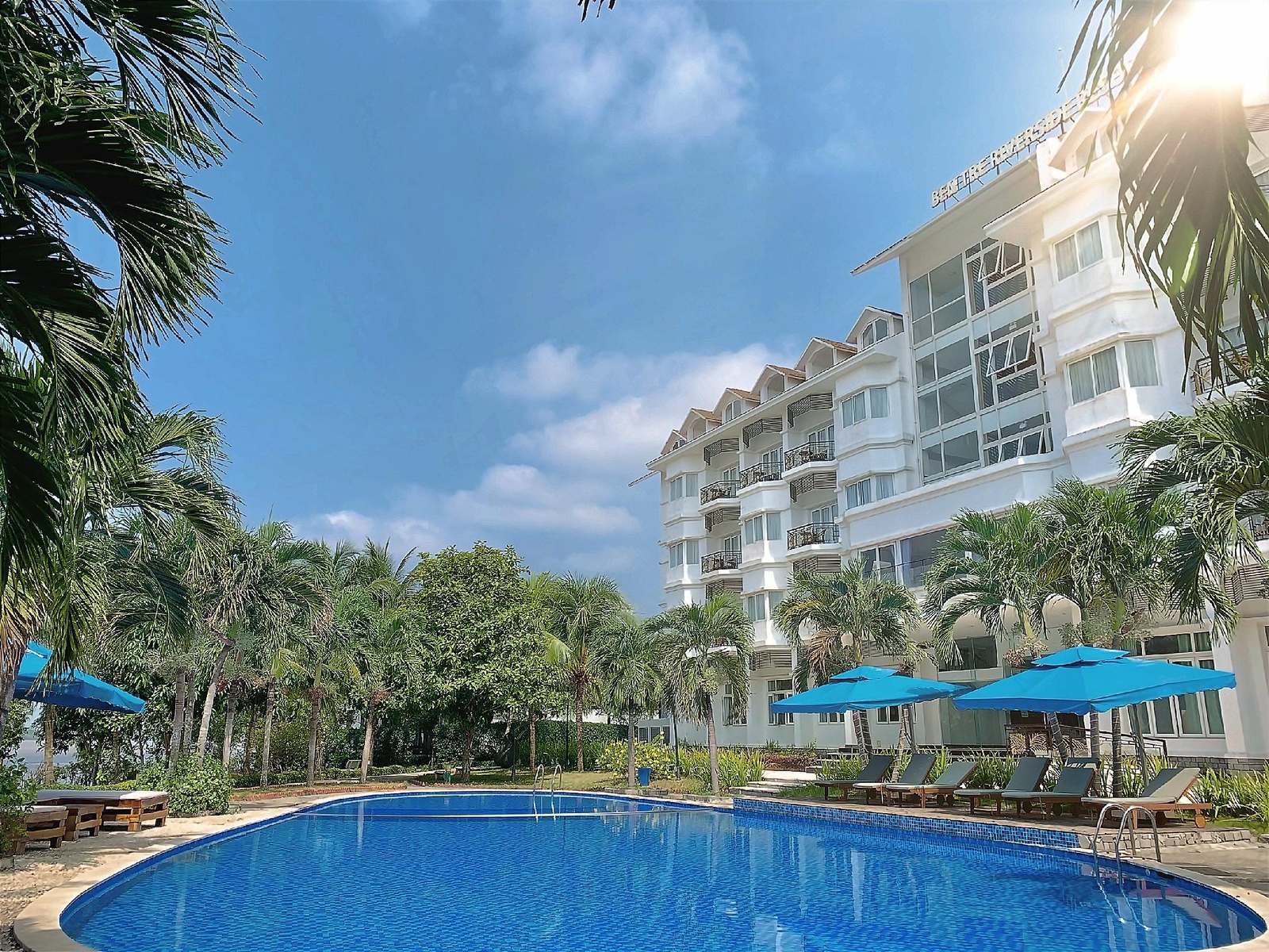 Curved outdoor pool with blue umbrellas and sun loungers in front of a white hotel block and palms