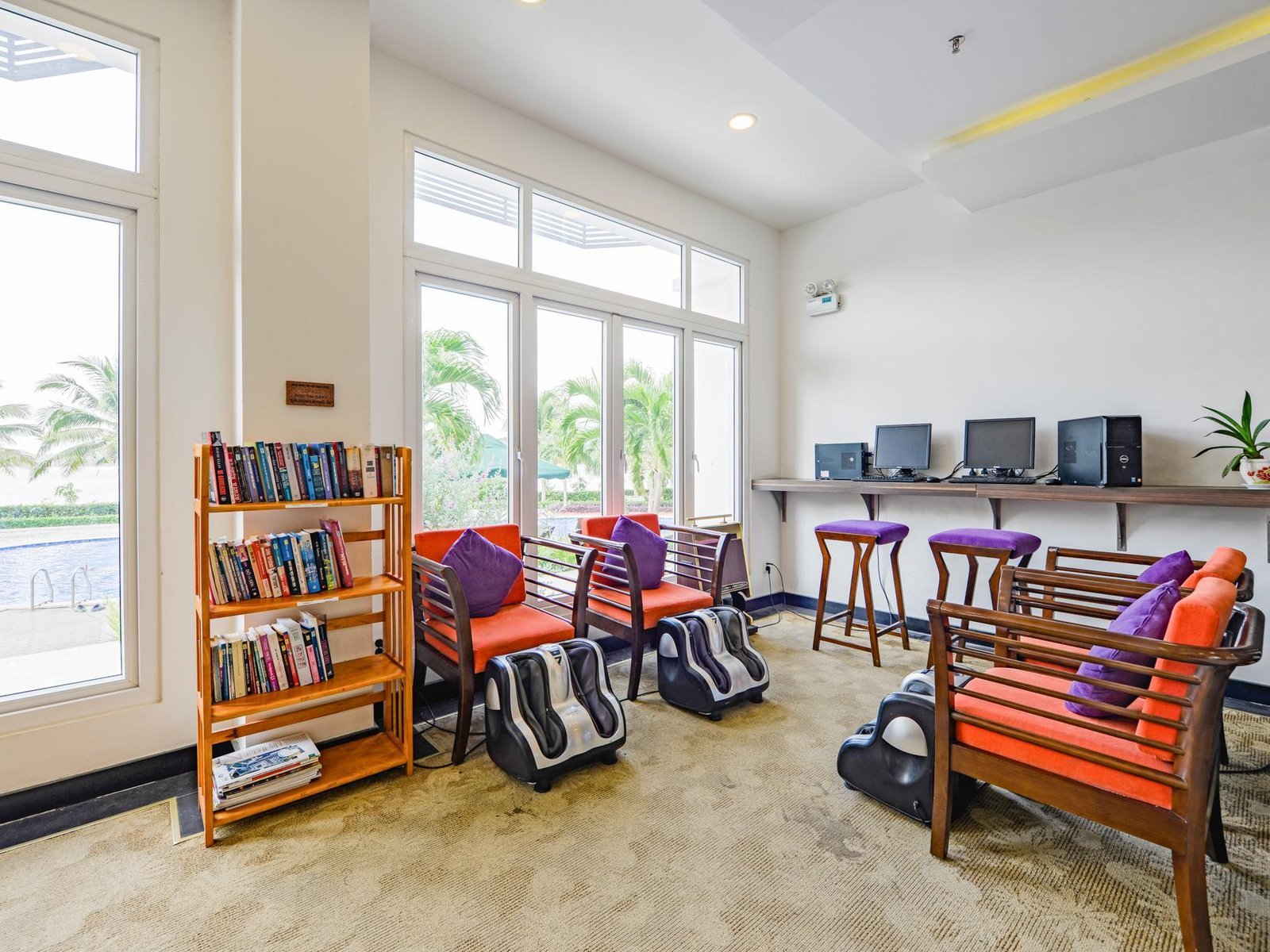 Lobby corner with bookshelves, orange armchairs, massage chairs and tall windows onto the garden