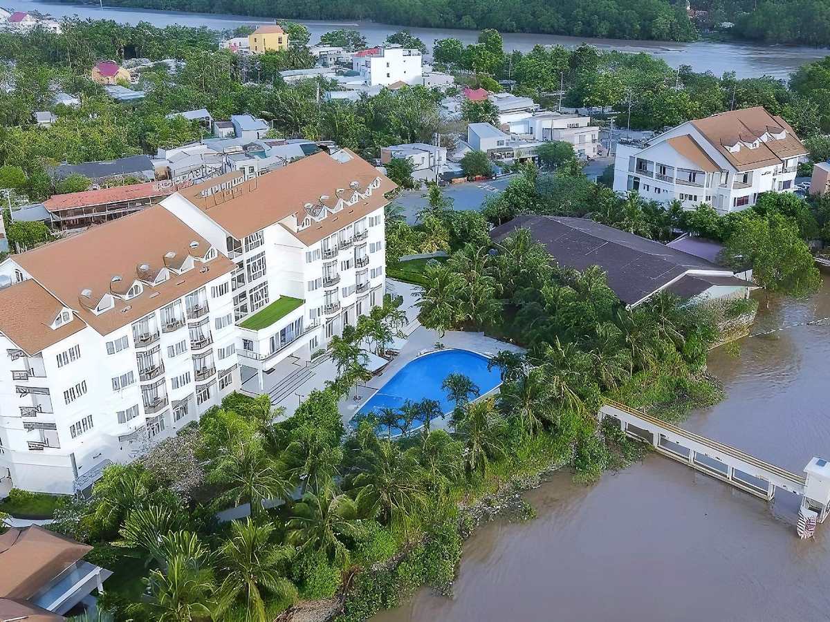 Aerial view of a white hotel block, a pool and a jetty on a wide brown river, with town houses behind