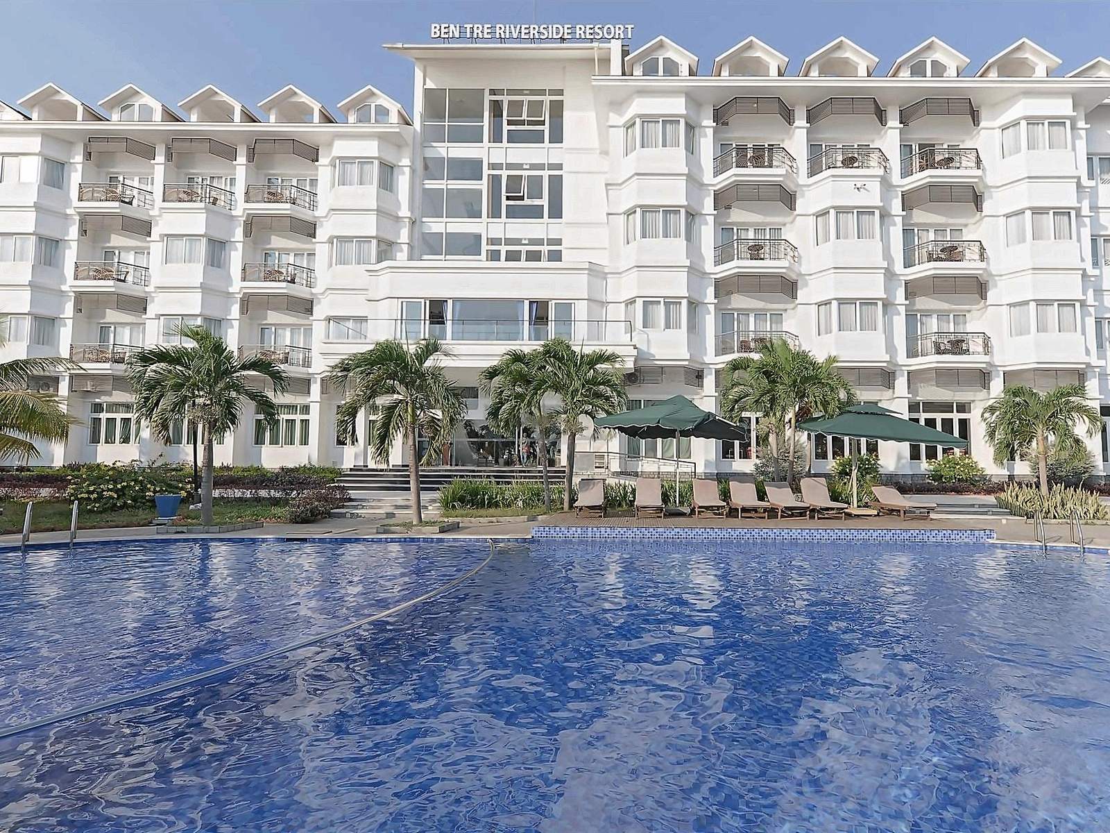 Bâtiment blanc à balcons se reflétant dans une piscine bleue, avec des palmiers au premier plan