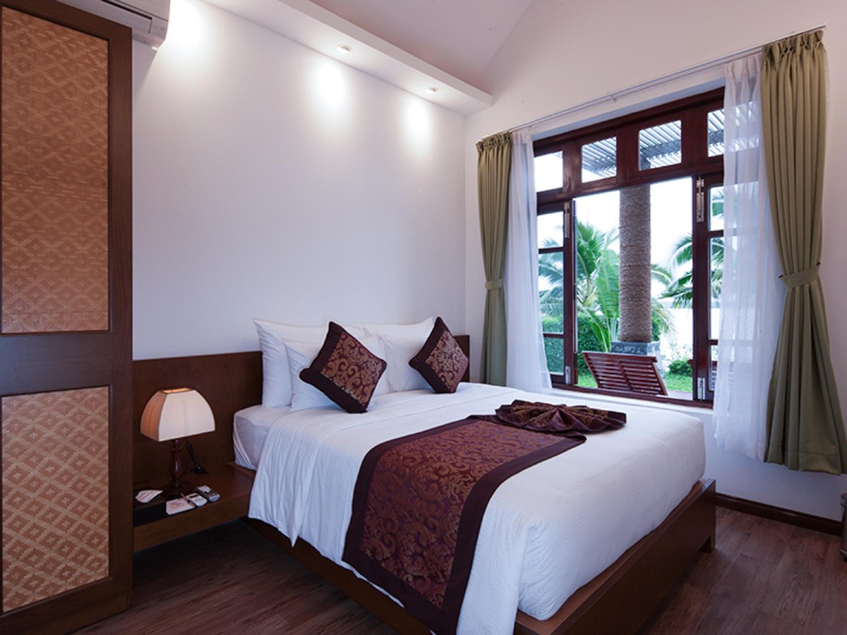 Bedroom with a dark wooden headboard, a vase of flowers and wood-framed windows looking onto palms
