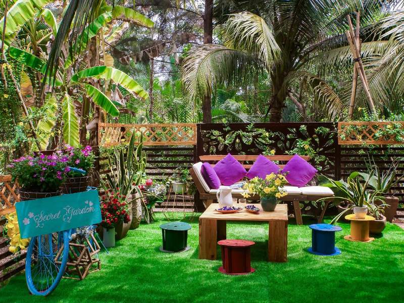 Sitzecke im Garten mit lila Kissen, Blumentöpfen und einem alten Fahrrad zwischen Bananenstauden und Kokospalmen
